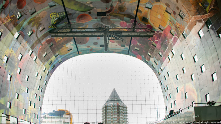 Markthal interior framing a pointed-roof tower through the tall gridded glass arch, with colorful mural imagery overhead.
