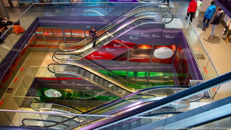 Several escalators crossing a multi-level atrium with bright red, green, purple, and blue wall graphics and a few visitors.