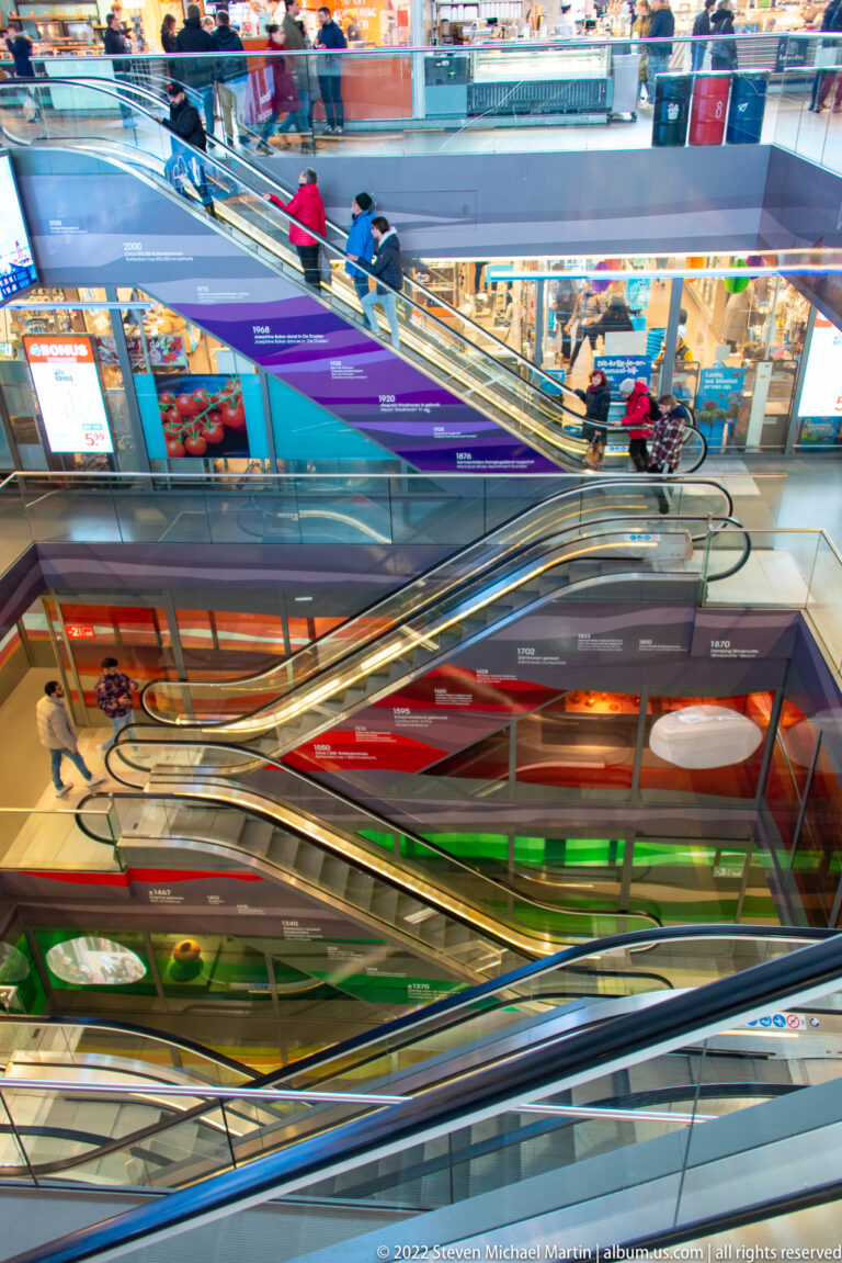 Side view of intersecting escalators with people above and bright red, purple, green, and blue wall graphics below.