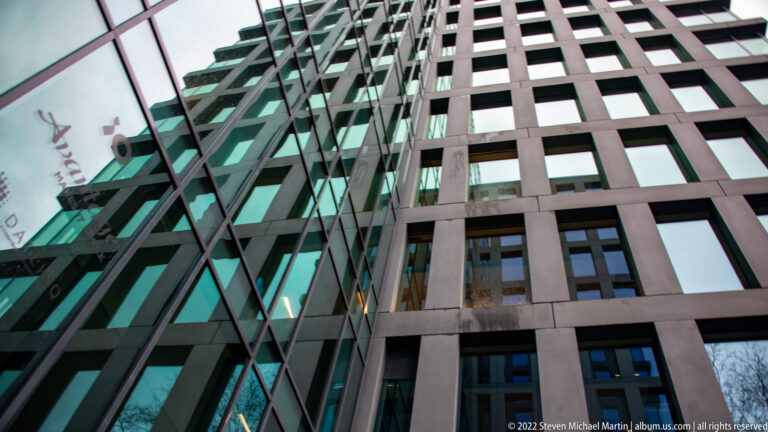Upward view between a green glass façade and a dark high-rise wall with a regular grid of recessed rectangular windows.
