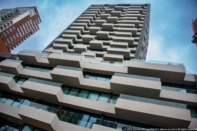 Modern residential tower seen from below, with many projecting pale balconies stacked irregularly against a blue-gray sky.