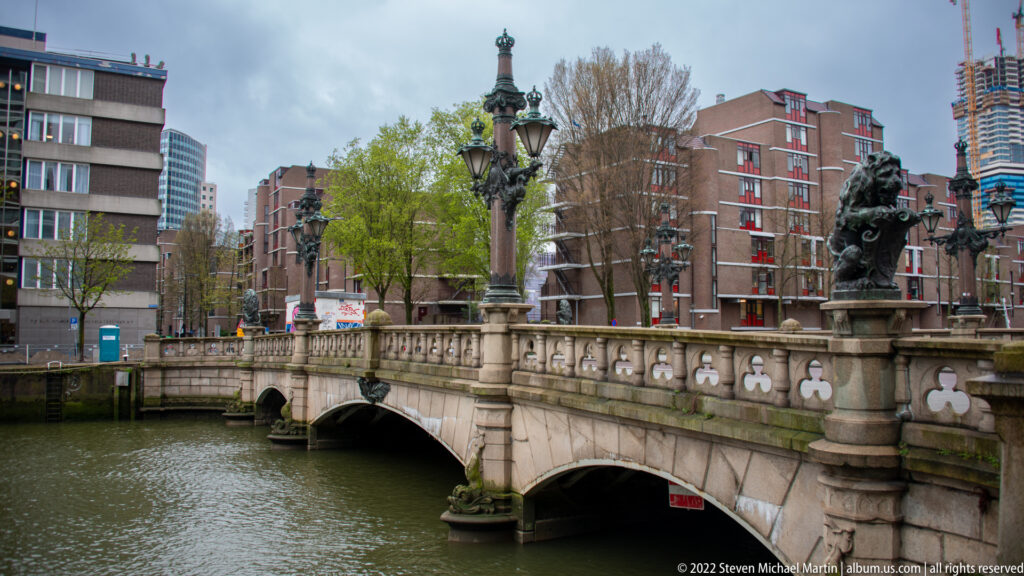 Stone bridge with carved balustrades, arches, ornate lamps, and sculptures crossing a harbor channel amid modern city buildings.