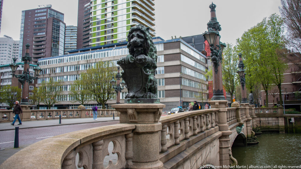 Dark sculpted bust mounted on an ornate stone bridge railing, with decorative lamps, trees, and modern buildings behind.