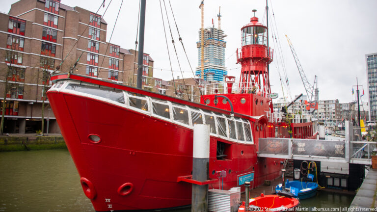 Large red lightship with a white-trimmed deckhouse and tall lantern tower moored in a Rotterdam harbor basin.