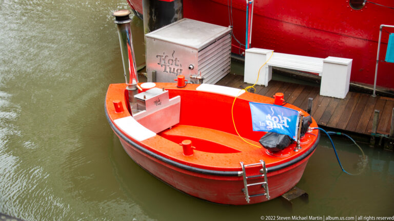 Small red open workboat tied beside a larger red vessel, with ladder, ropes, bench, and blue cover visible from above.