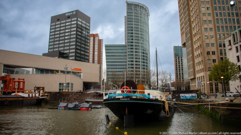 Historic dark-hulled vessel moored in a narrow city harbor, surrounded by modern office and residential towers.