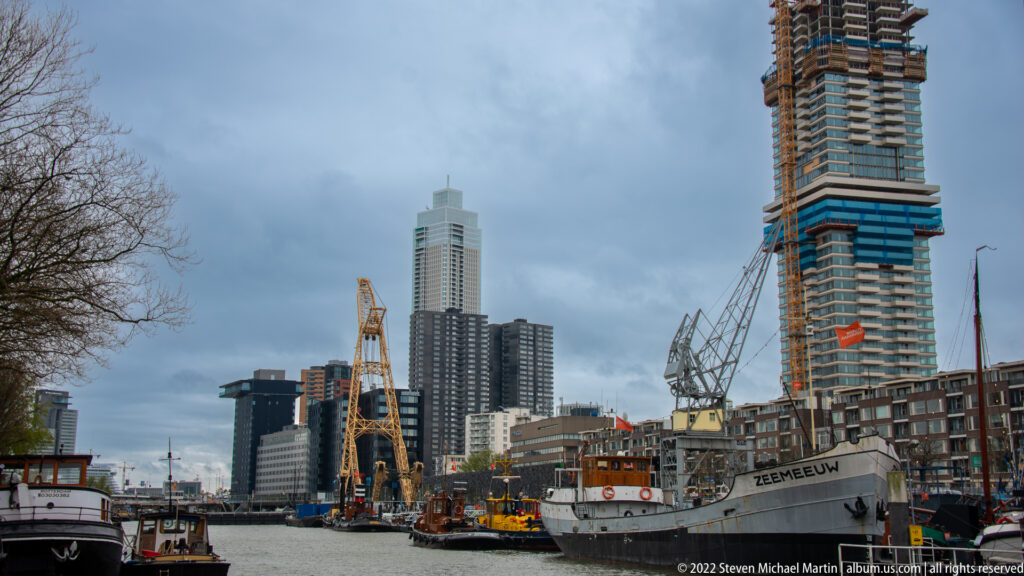 Historic vessels and cranes in a Rotterdam harbor, with the vessel Zeemeeuw at right and modern high-rise towers behind.