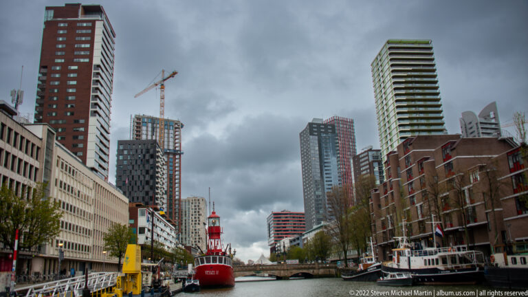 Long view down a Rotterdam harbor basin with a red lightship centered between high-rise towers, cranes, and waterfront buildings.