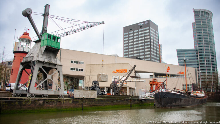 Historic metal harbor crane with elevated cab beside museum vessels, with the Maritime Museum and office towers behind.