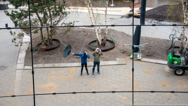 Two visitors gesturing upward, reflected in mirrored panels with birch trees, bicycles, and a service vehicle behind them.