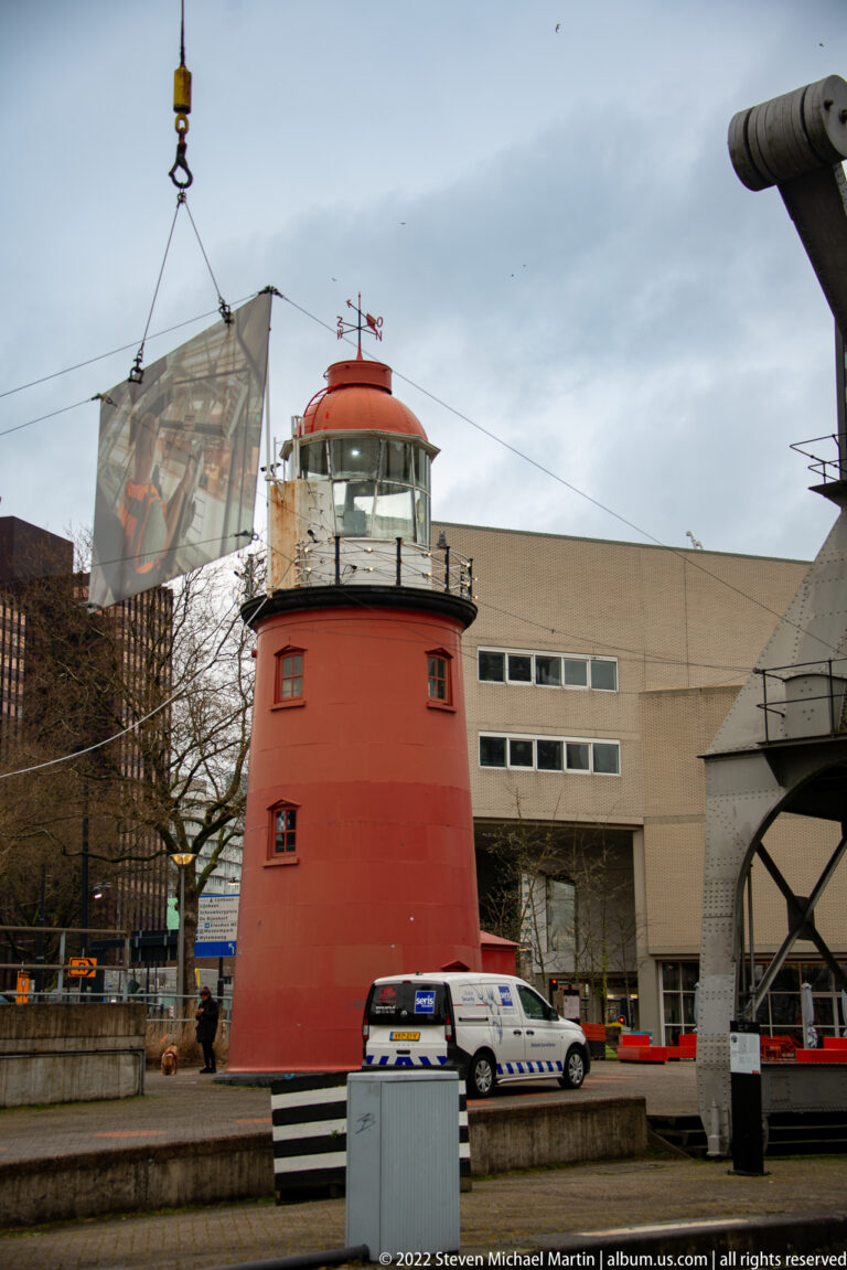 Red cylindrical harbor-side tower with glazed top, striped base, service vehicles, and modern waterfront buildings behind.