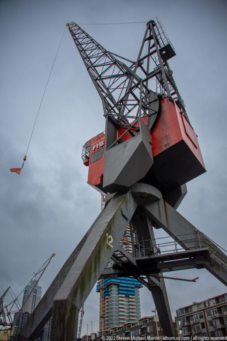 Large red-and-gray lattice-boom harbor crane viewed from below against a cloudy sky.