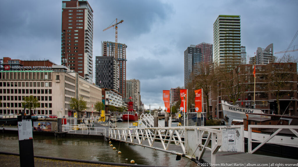 Pedestrian gangway over a harbor basin with offices, high-rise towers, and construction cranes in central Rotterdam.