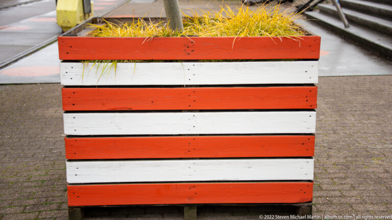 Rectangular street planter painted in alternating red and white horizontal stripes with grasses growing from the top.