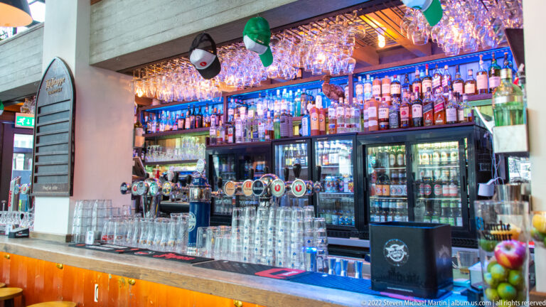 Pub bar with rows of bottles, beer taps, hanging glassware, refrigerators, and blue backlighting.
