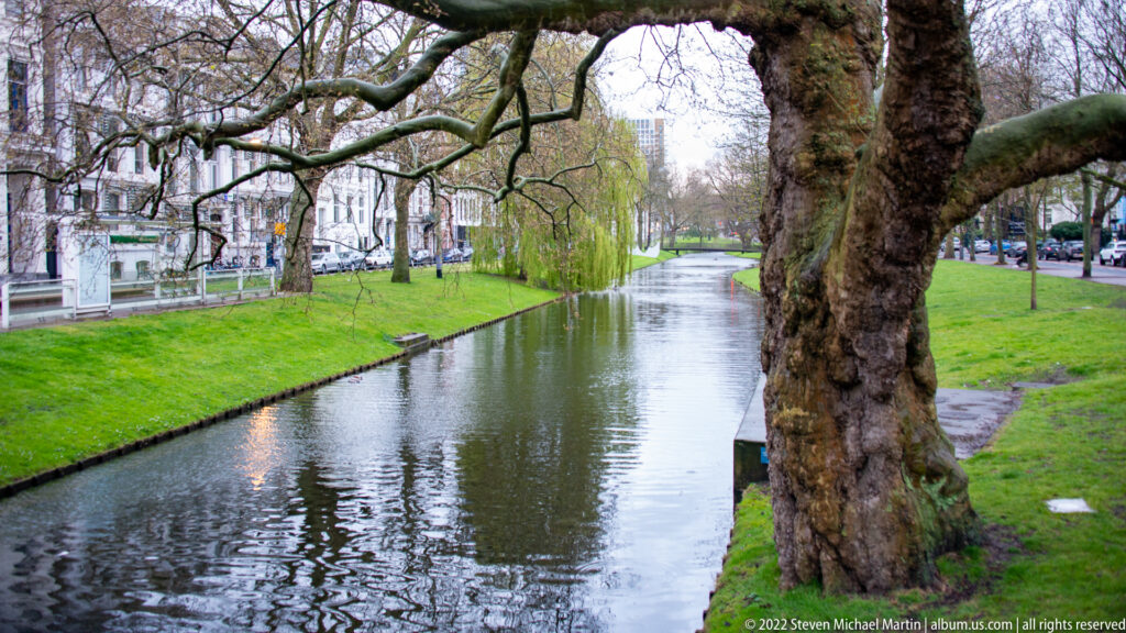 Narrow canal bordered by grass and mature trees, with pale urban buildings along the left bank.