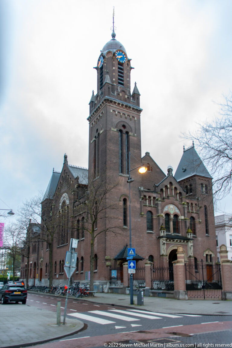 Large historic brick church with square bell tower, arched windows, street signs, and bicycles at a Rotterdam intersection.
