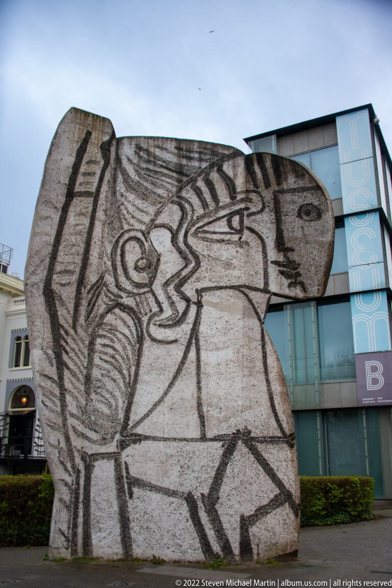 Large irregular concrete sculpture with black incised lines forming a stylized human figure beside modern buildings.