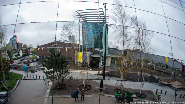 Wide mirrored reflection of Museumpark with buildings, birch trees, pathways, construction, and two visitors.