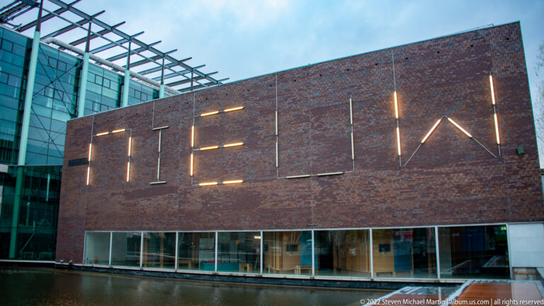 Dark brick cultural building with large illuminated linear letters above a glazed ground floor beside water.