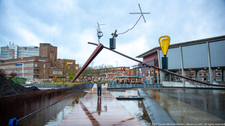 Angular outdoor sculpture with red, black, and yellow elements over a wet walkway in Museumpark.