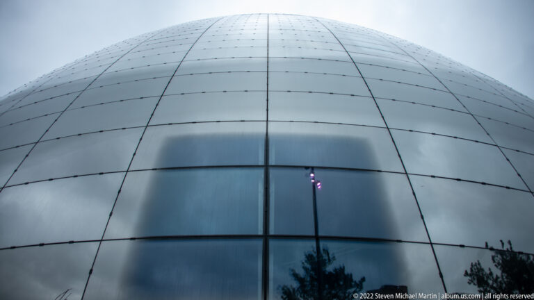 Close upward view of Depot Boijmans Van Beuningen’s curved mirrored panels with faint reflections of trees and a lamp.