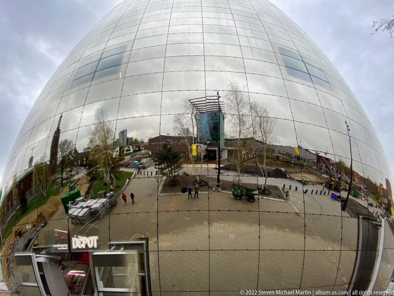 Wide view of Depot Boijmans Van Beuningen’s curved mirrored façade reflecting Museumpark plaza, people, trees, and buildings.