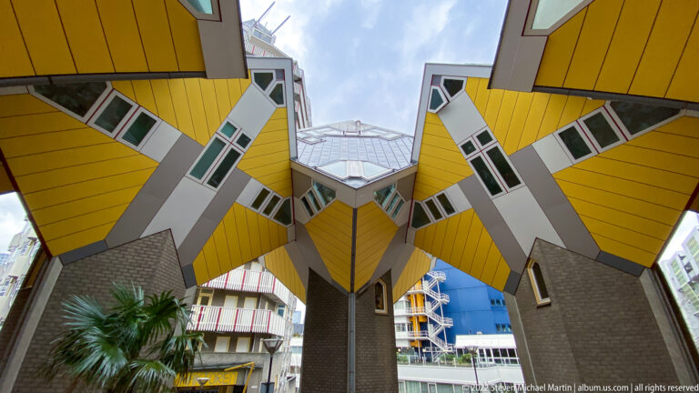 Upward view beneath Rotterdam’s yellow Cube Houses showing tilted façades, angular windows, and gray structural supports.