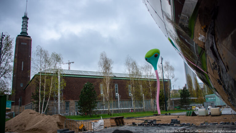 Brick church and tower behind birch trees, construction works, and a tall green-and-pink sculptural form.