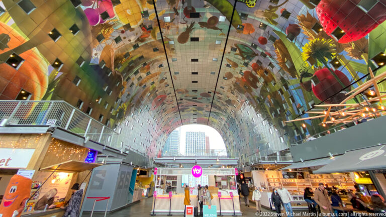 Interior of Rotterdam Markthal with food stalls below the immense colorful arched ceiling mural and glazed end wall.