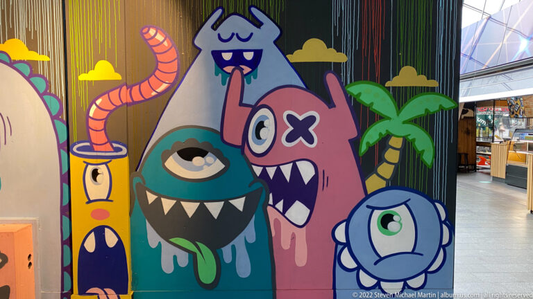 Colorful cartoon monster mural with one-eyed creatures, fangs, horns, drips, and palm-tree motifs inside a market setting.