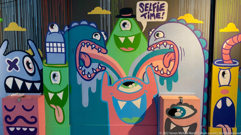 Colorful cartoon monster mural reading SELFIE TIME, with one-eyed, horned, and toothy characters packed across the wall.