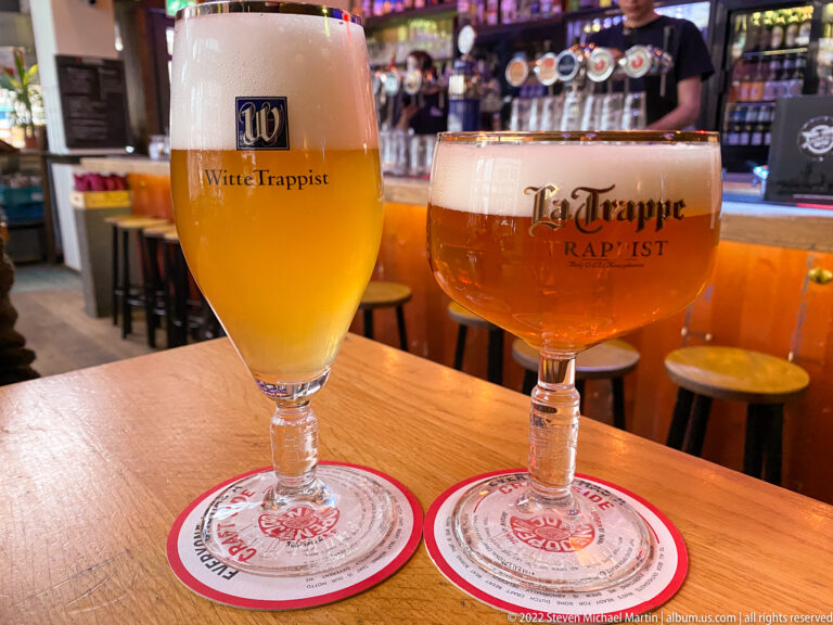 Two stemmed Trappist beer glasses, Witte Trappist and La Trappe Trappist, on coasters at a bar table.