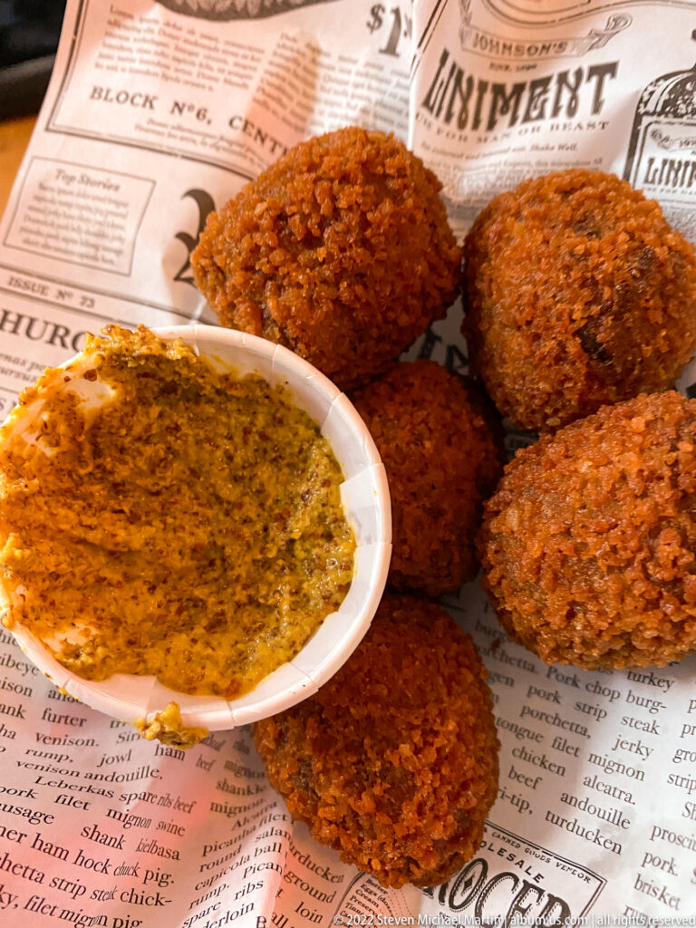 Several round fried bitterballen on printed paper beside a small dish of coarse mustard.