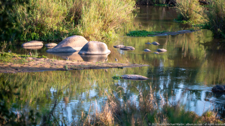 Calm river pool with rounded boulders, low grassy banks, and tree reflections on still water.