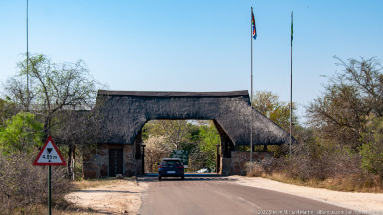 Thatched stone gateway spanning a paved Kruger road, with vehicles, flagpoles, and dry woodland on both sides.