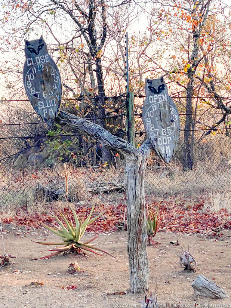 Rustic wooden posts holding shield-shaped CLOSED and OPEN signs beside aloes and a fence.
