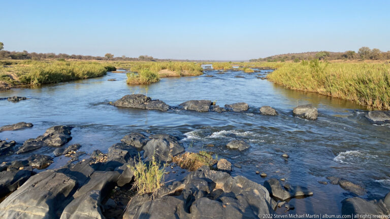 Shallow river flowing around dark rocks and low reed-covered islands under a clear sky.