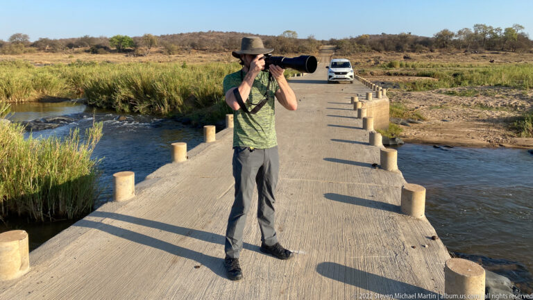 Photographer with a long telephoto lens standing on a concrete low-water bridge over a shallow river.