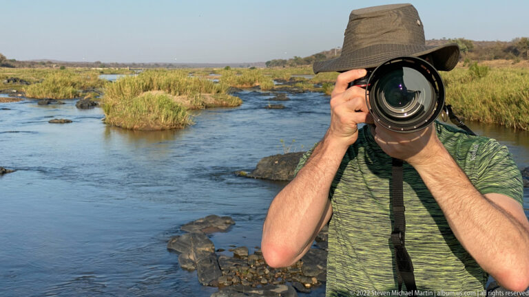 Close view of a photographer aiming a large camera lens, with a shallow river and reeds behind.