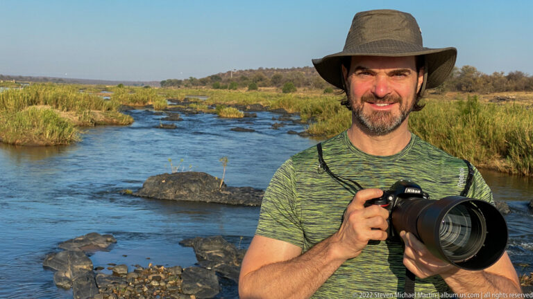 Traveler in a brimmed hat holds a telephoto camera beside a rocky river with reeds and low woodland.