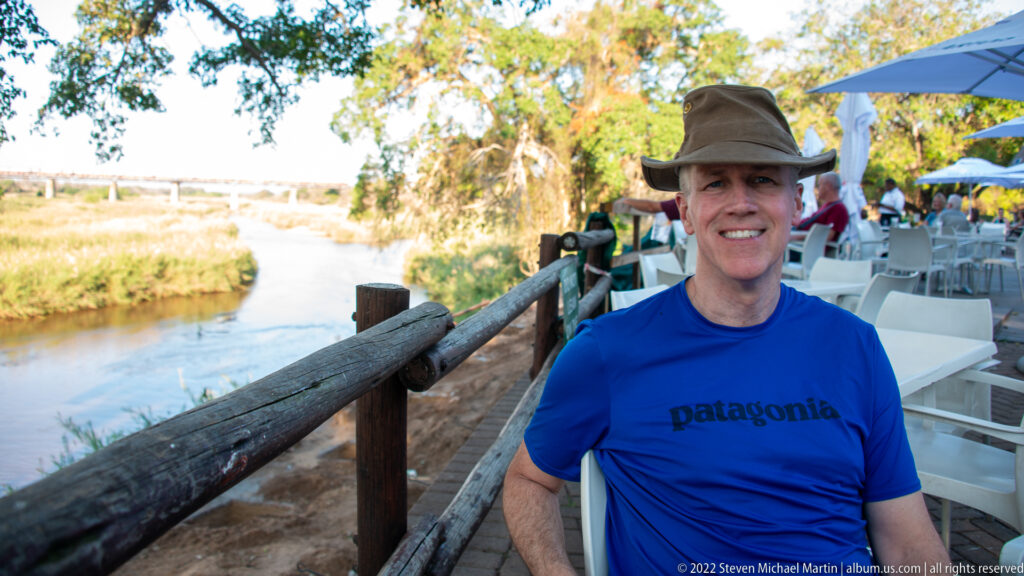 Traveler in a brimmed hat and blue shirt seated beside a river and outdoor dining area.