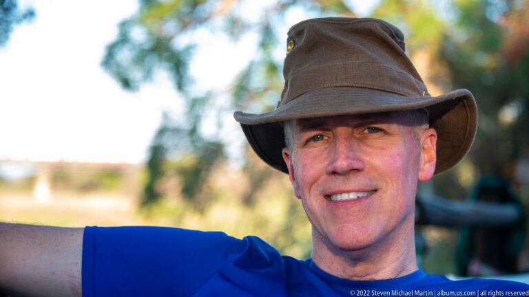 Smiling traveler in a broad-brimmed hat and blue shirt, with a softly blurred outdoor background.