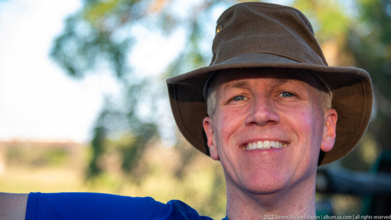 Tight smiling portrait of a traveler wearing a soft brown safari hat against blurred greenery.