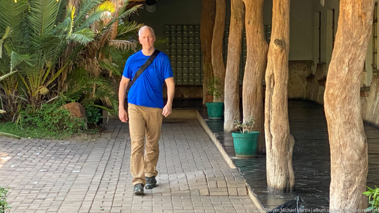 Traveler in a blue shirt walks along a paved path beside thick timber posts and leafy landscaping.