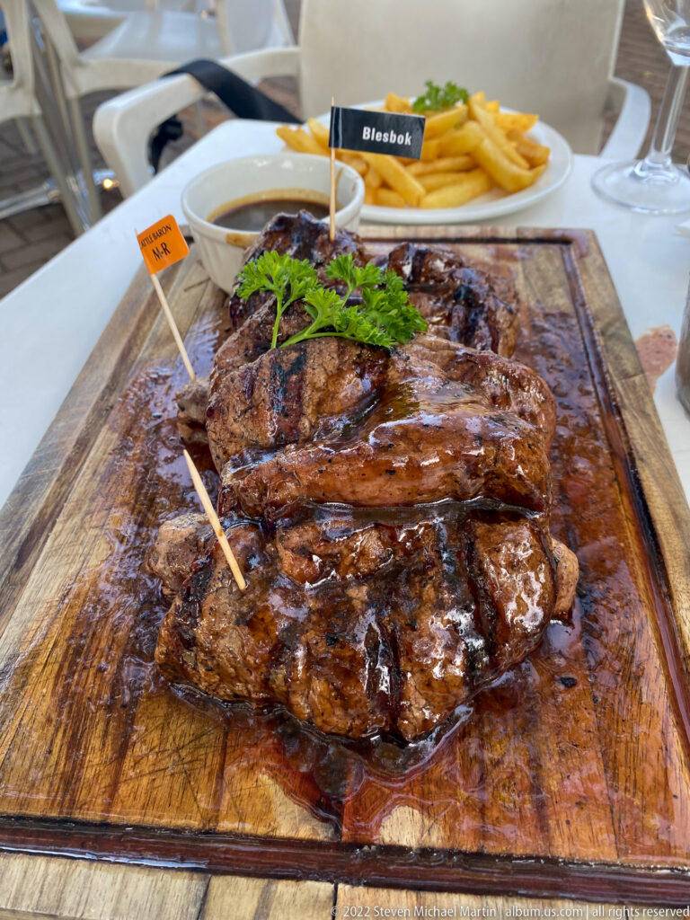 Several grilled meat portions on a wooden serving board with fries, greens, and small label flags.
