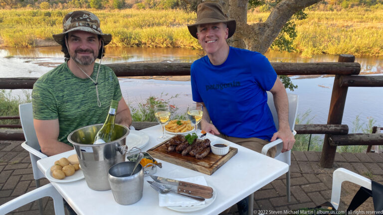 Two travelers seated at an outdoor table with grilled food and wine beside a river at sunset.