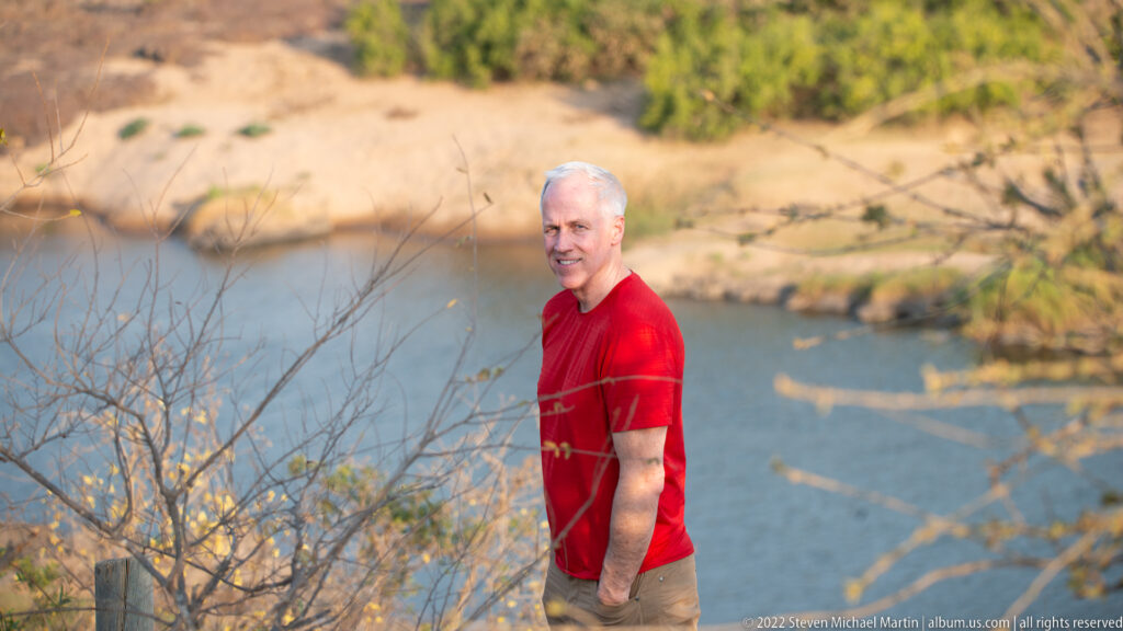 Visitor in a red shirt standing above the Olifants River with water and a pale opposite bank behind.