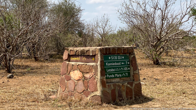 Stone Kruger National Park directional marker with painted destination panels and arrows beside a dry bushveld road.