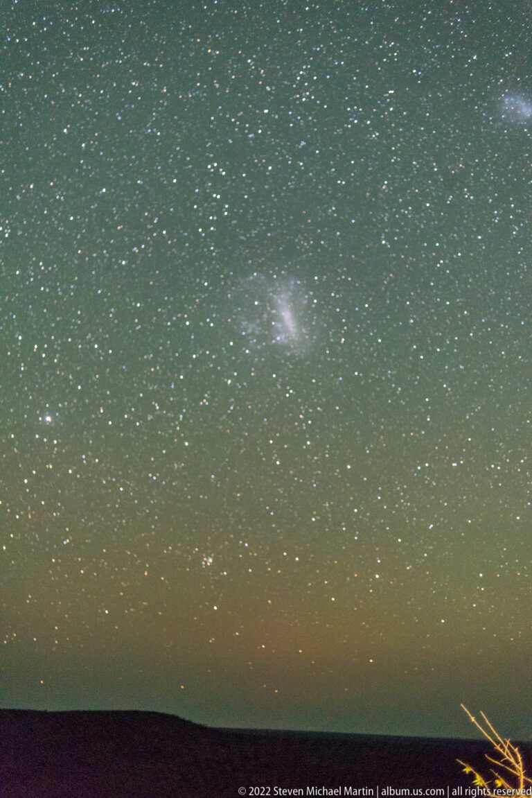 Dense star field above a dark horizon with a bright diffuse cloudlike patch near the center.